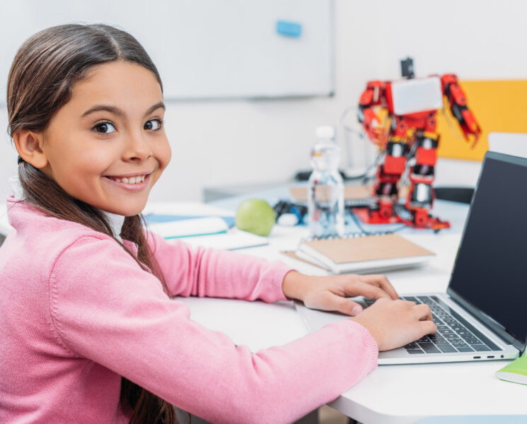 adorable schoolgirl sitting at table with robot model, looking at camera and using laptop with blank screen during STEM lesson