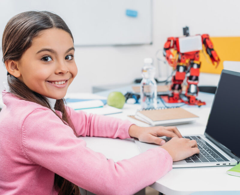 adorable schoolgirl sitting at table with robot model, looking at camera and using laptop with blank screen during STEM lesson